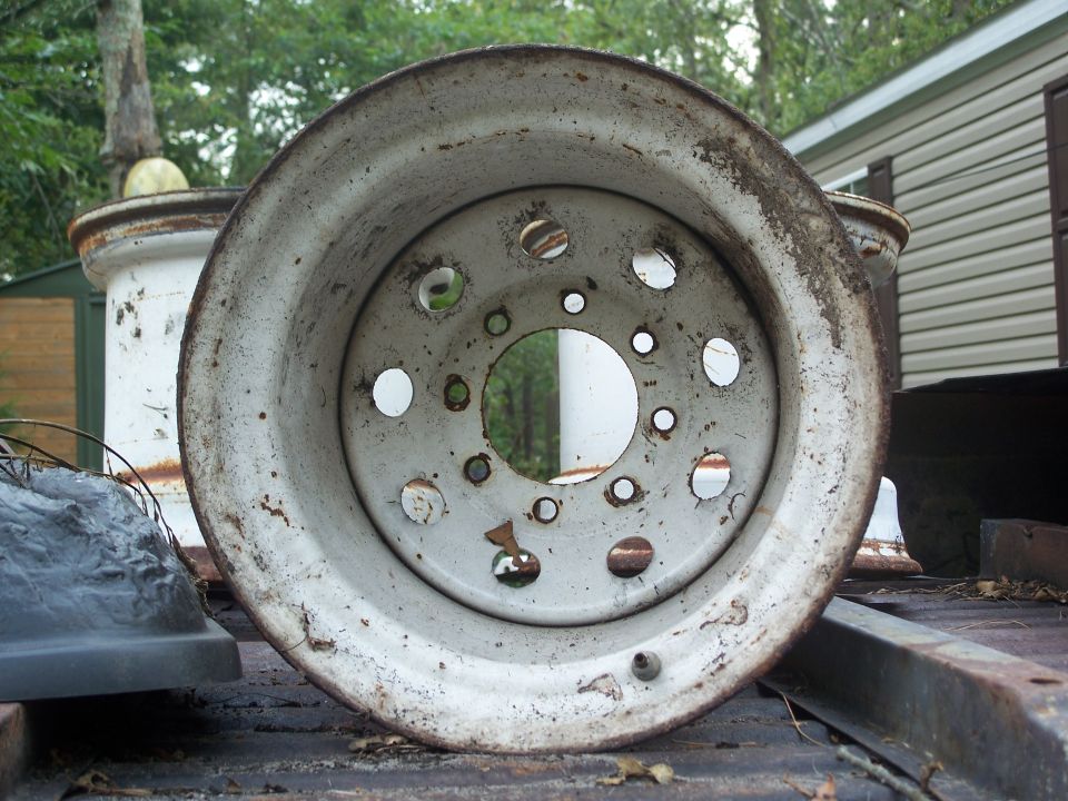 Close-up of a rusty white wheel rim with holes, with a building in the background.