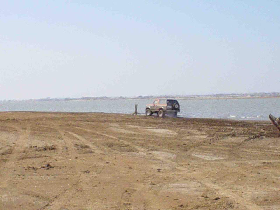 Tan 1980s Ford Bronco with a black spare tire carrier parked on a dirt shore near water