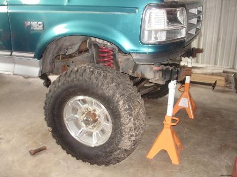 1996 Ford F-150 - The front driver's side of a teal 1990s Ford F-150 pickup truck is shown, lifted with a large off-road tire. The front end is supported by…