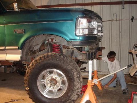 1996 Ford F-150 - A person works on the front suspension of a teal green Ford F-150 pickup truck that is raised on jack stands. The front tire and part of th…