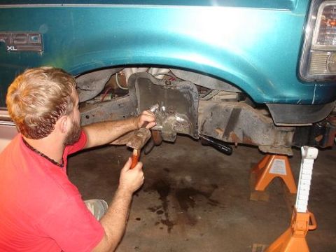 1996 Ford F-150 - A man in a red shirt uses a hammer and chisel to work on the rusted frame of a teal Ford F-150 pickup truck, which is supported by jack sta…