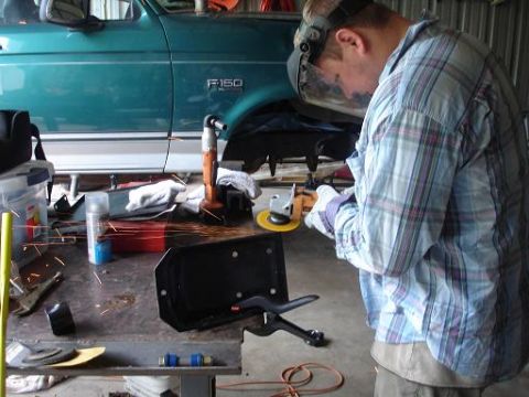 1996 Ford F-150 - A person wearing a welding mask and plaid shirt grinds a metal part on a workbench next to a teal Ford F-150 pickup truck.