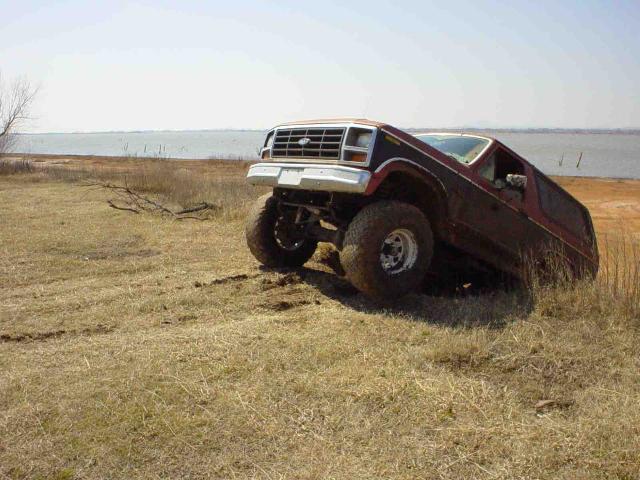 1980s Ford Bronco with lifted suspension and large tires tilted on grassy terrain near water