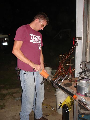 1996 Ford F-150 - A man wearing safety glasses and a maroon t-shirt is using an angle grinder to cut a metal pipe clamped to a workbench. Sparks are flying f…
