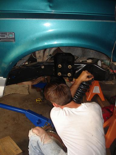 1996 Ford F-150 - A person works on the suspension of a teal Ford truck, with a coil spring held by a jack stand visible.