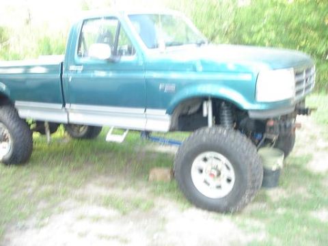 1996 Ford F-150 - A teal Ford F-150 pickup truck is shown from the front 3/4 angle, with a lifted suspension and large off-road tires. A white bucket is visi…