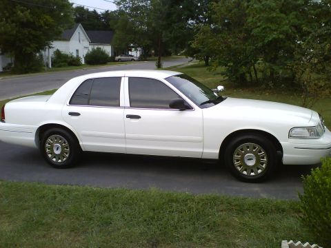 2003 Ford Crown Victoria - A white 2003 Ford Crown Victoria sedan is parked on a paved driveway in front of a grassy area and trees. The car is viewed from the side a…