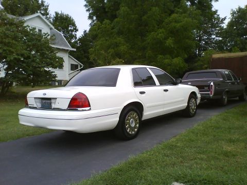 2003 Ford Crown Victoria - A white 2003 Ford Crown Victoria is parked on a driveway next to a dark-colored sedan. A white house and trees are visible in the backgroun…