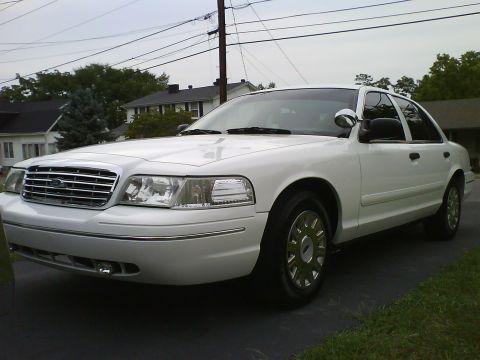 2003 Ford Crown Victoria - A white 2003 Ford Crown Victoria sedan is parked in a driveway with houses and trees in the background. The front of the car is angled towa…