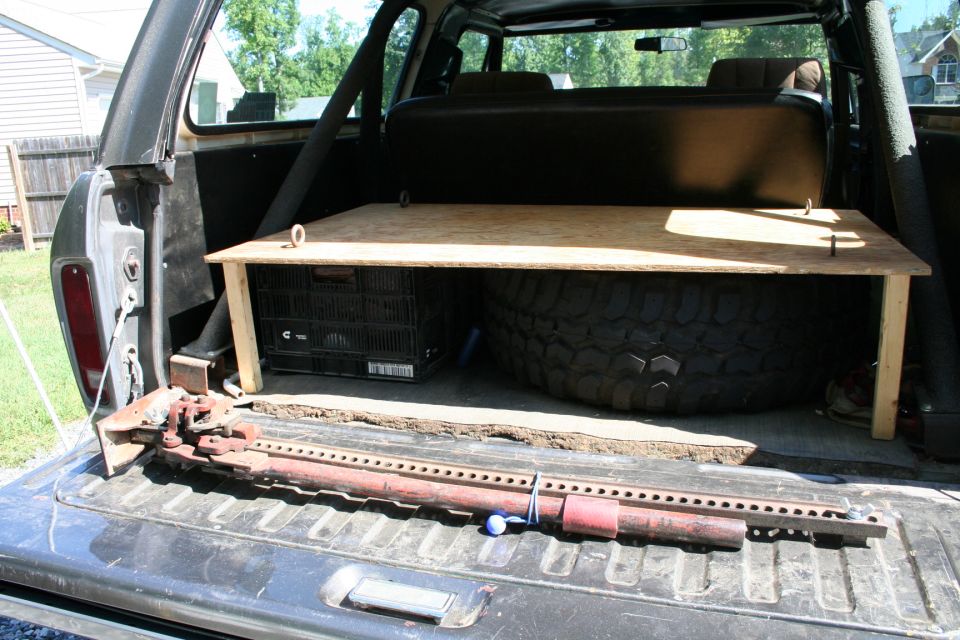 Open tailgate of a dark SUV showing a wooden platform, spare tire, crate, and hi-lift jack.