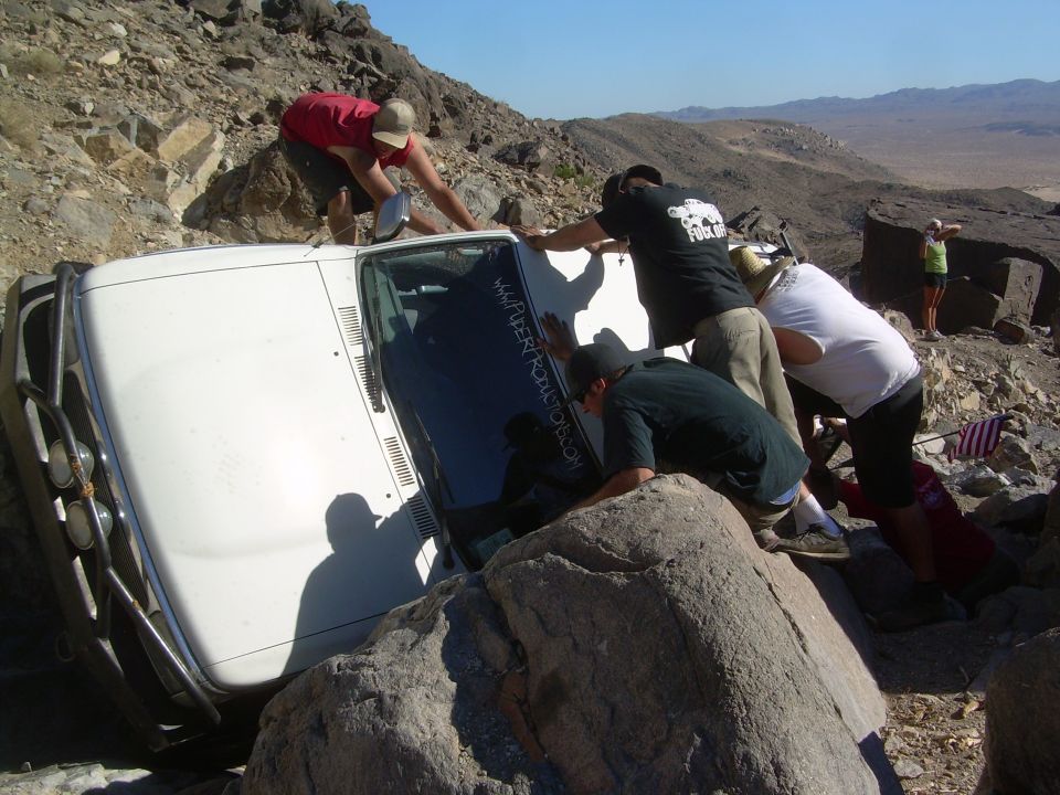 Five people righting a white 4Runner rolled onto its side among desert boulders