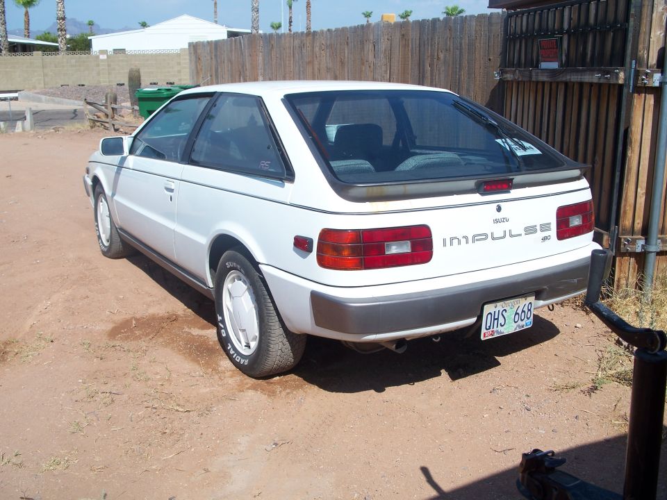 White Isuzu Impulse hatchback, rear 3/4 view, parked on dirt by a wooden fence.