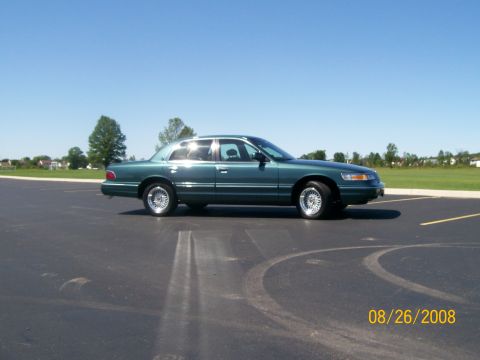 1995 Mercury Grand Marquis - A dark green sedan is parked on an asphalt surface with yellow lines. The car is viewed from the side, with the front wheel turned slightly.
