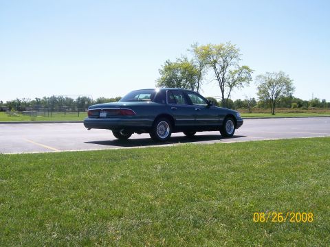 1995 Mercury Grand Marquis - A dark green 1995 Mercury Grand Marquis sedan is parked in a paved lot next to a grassy area. The car is viewed from the rear quarter angle…