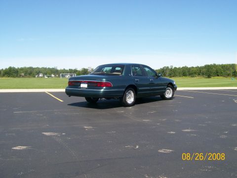 1995 Mercury Grand Marquis - A dark green sedan is parked in a black asphalt parking lot with a grassy field and trees in the background under a clear blue sky. The rea…