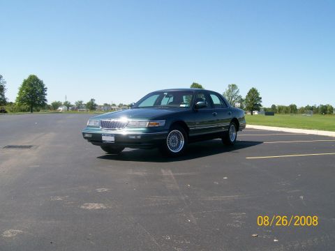 1995 Mercury Grand Marquis - A dark green sedan is parked in an asphalt lot on a sunny day. The vehicle is angled towards the camera, showing its front and side profile.