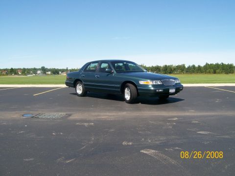 1995 Mercury Grand Marquis - A dark green 1995 Mercury Grand Marquis sedan is parked in an empty asphalt lot under a clear blue sky. The car is viewed from the front th…