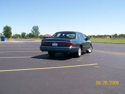 1995 Mercury Grand Marquis - A dark green sedan is parked in an empty asphalt parking lot under a clear blue sky. The rear driver's side of the car is visible, along wi…