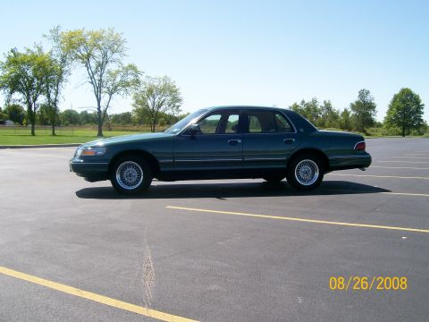 1995 Mercury Grand Marquis - A dark green sedan is parked in an empty asphalt lot with yellow parking lines. The car is viewed from the side profile under a clear blue…