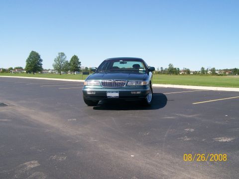 1995 Mercury Grand Marquis - A dark green sedan is parked in an empty asphalt parking lot on a clear day. The front of the car is facing the camera, with a grassy field…