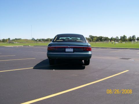 1995 Mercury Grand Marquis - The rear of a dark green sedan parked in an empty asphalt parking lot on a sunny day. Yellow lines mark parking spaces and a date stamp rea…