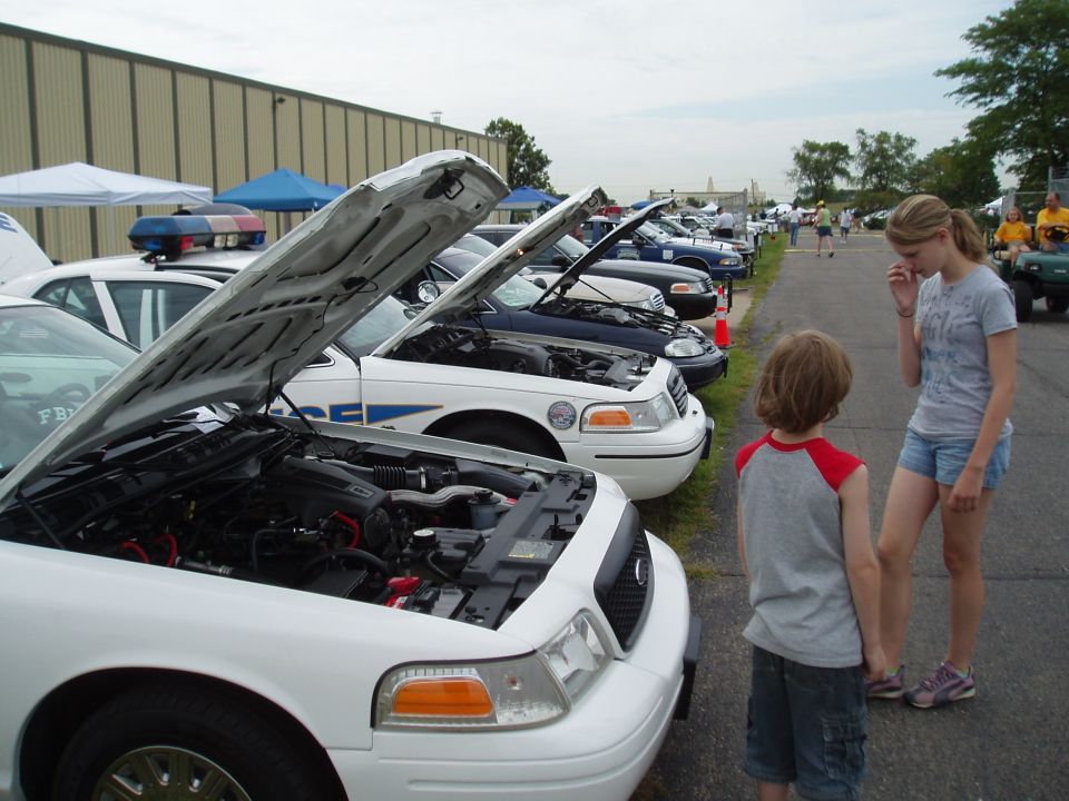 Several Ford Crown Victoria police cars with hoods open, parked in a lot. Two people stand in foreground.
