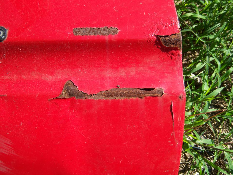 Red vehicle panel with rust and peeling paint, outdoors with grass.