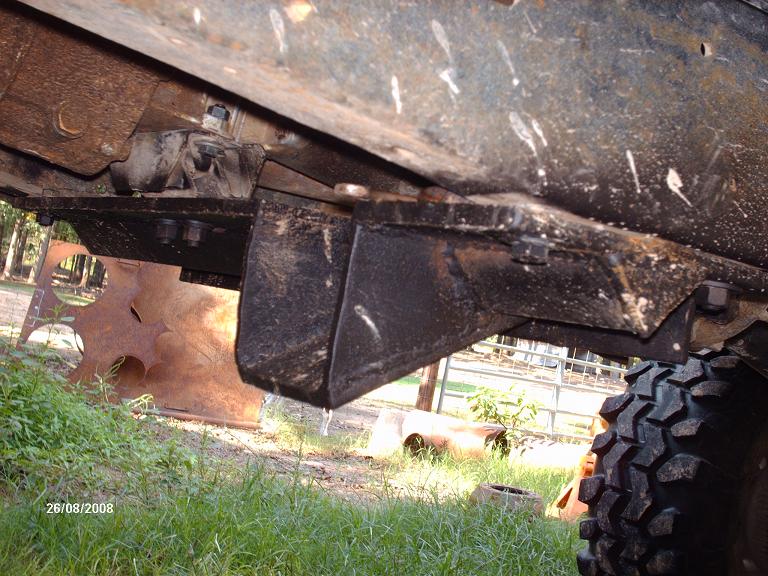 Undercarriage of a vehicle showing suspension and frame components, with grass and trees in the background.