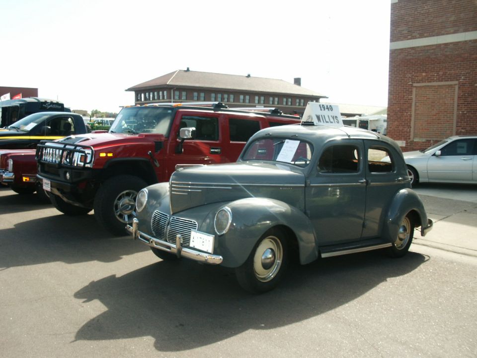 1940 Willys sedan and red Hummer H2 parked in front of a brick building.