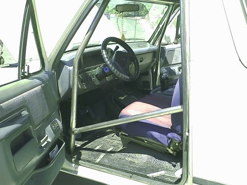 1990 Ford Bronco - Interior view of a 1990 Ford Bronco, showing the driver's side door, dashboard, steering wheel, and roll cage. The vehicle has a blue and p…