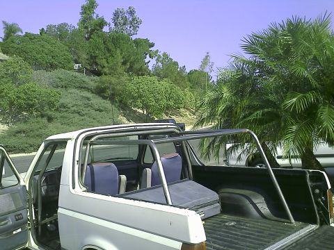 1990 Ford Bronco - The rear passenger side of a white 1990 Ford Bronco is shown with a roll cage installed in the interior and bed. The vehicle is parked outd…