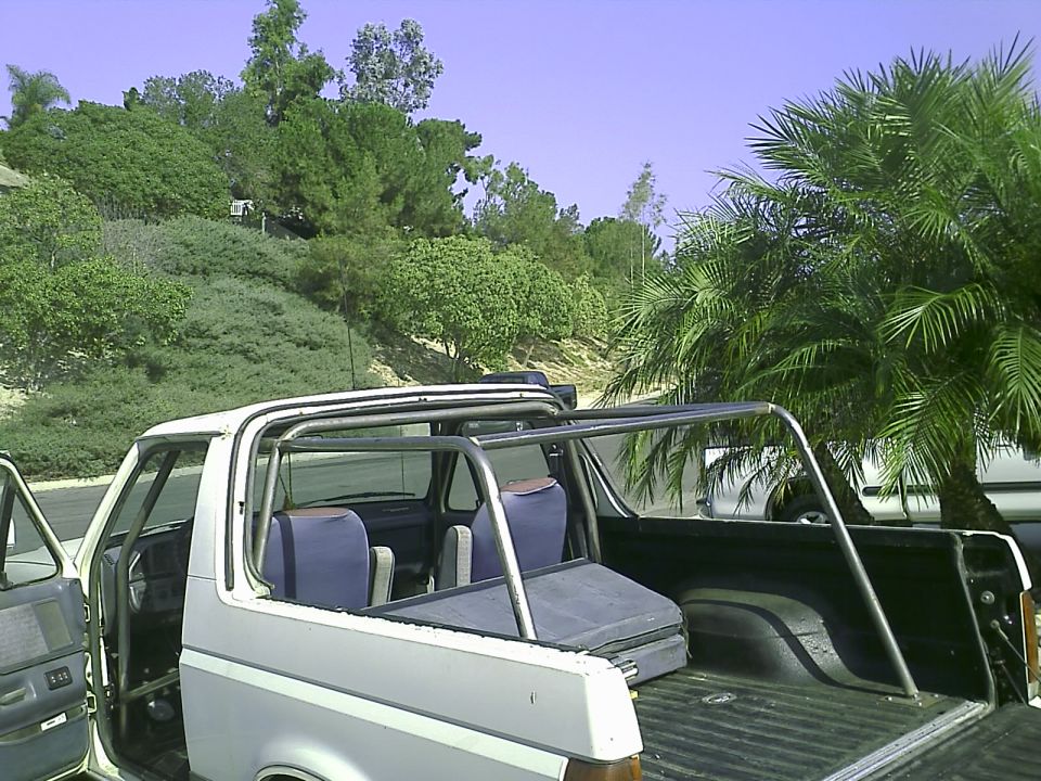 White 1990 Ford Bronco with roll cage, parked outdoors with hills and palm trees