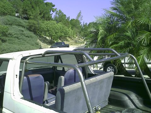 1990 Ford Bronco - Interior view of a white Ford Bronco with a custom roll cage and blue seats. A dark pickup truck is visible in the background on a dirt roa…
