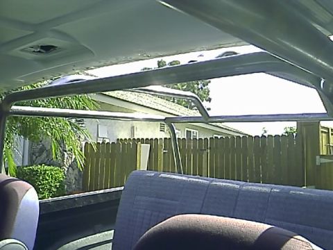 1990 Ford Bronco - Interior view of a roll cage installed in a vehicle, looking towards the rear seats and out through the open top at a wooden fence and tree…