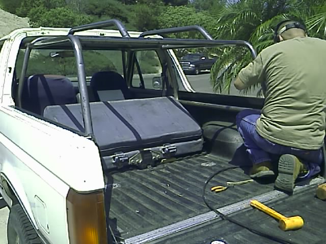 Man working on roll cage of white 1990 Ford Bronco convertible with toolbox in bed.
