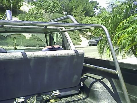 1990 Ford Bronco - The interior of a pickup truck bed shows a roll cage and the back of the seats. A dark-colored SUV is visible in the background.
