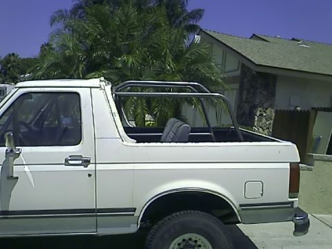 1990 Ford Bronco - The side view of a white Ford Bronco with a roll cage installed in the bed. The vehicle is parked outdoors with palm trees and a house in t…