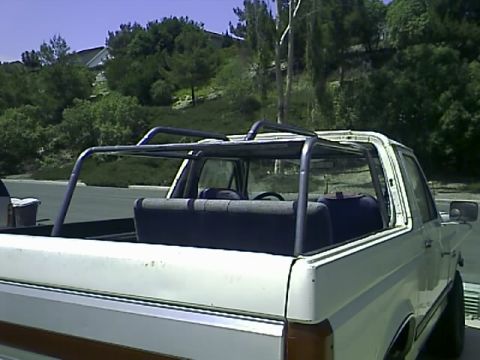 1990 Ford Bronco - White pickup truck bed with an installed roll bar and folding rear bench seat, hills in background.