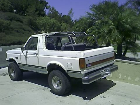 1990 Ford Bronco - The rear three-quarter view of a white Ford Bronco with a roll cage installed in the open bed area. The vehicle is parked on a concrete sur…