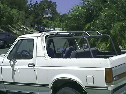 1990 Ford Bronco - The side view of a white Ford Bronco with a custom roll cage installed in the open-top rear. The vehicle is parked on a street with palm tr…