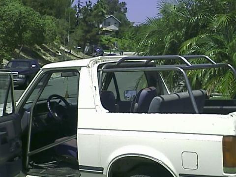 1990 Ford Bronco - Side view of a white Ford Bronco with its hardtop removed showing the roll cage and rear seat, parked on a hillside street near palms.