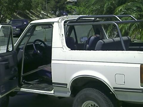 1990 Ford Bronco - Driver's-side view of a white full-size Ford SUV with its removable rear hardtop taken off, exposing the roll bar, rear bench seat and open…