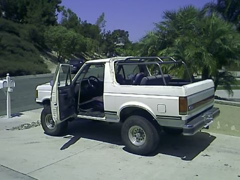 1990 Ford Bronco - A white 1990 Ford Bronco with its doors open is parked on a paved driveway. The vehicle has a roll cage installed in the rear and is surrou…