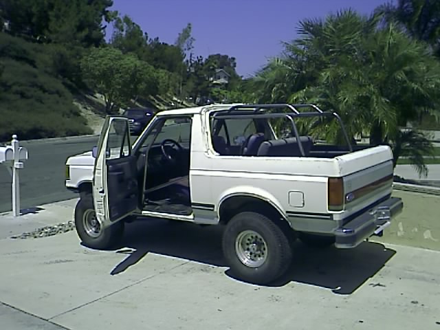 White 1990 Ford Bronco with open doors and a roll cage, parked in a driveway.