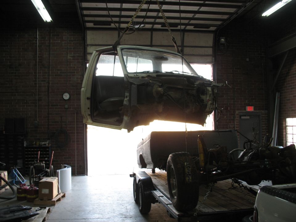 Disassembled vehicle cab suspended by chains, chassis on trailer in garage.