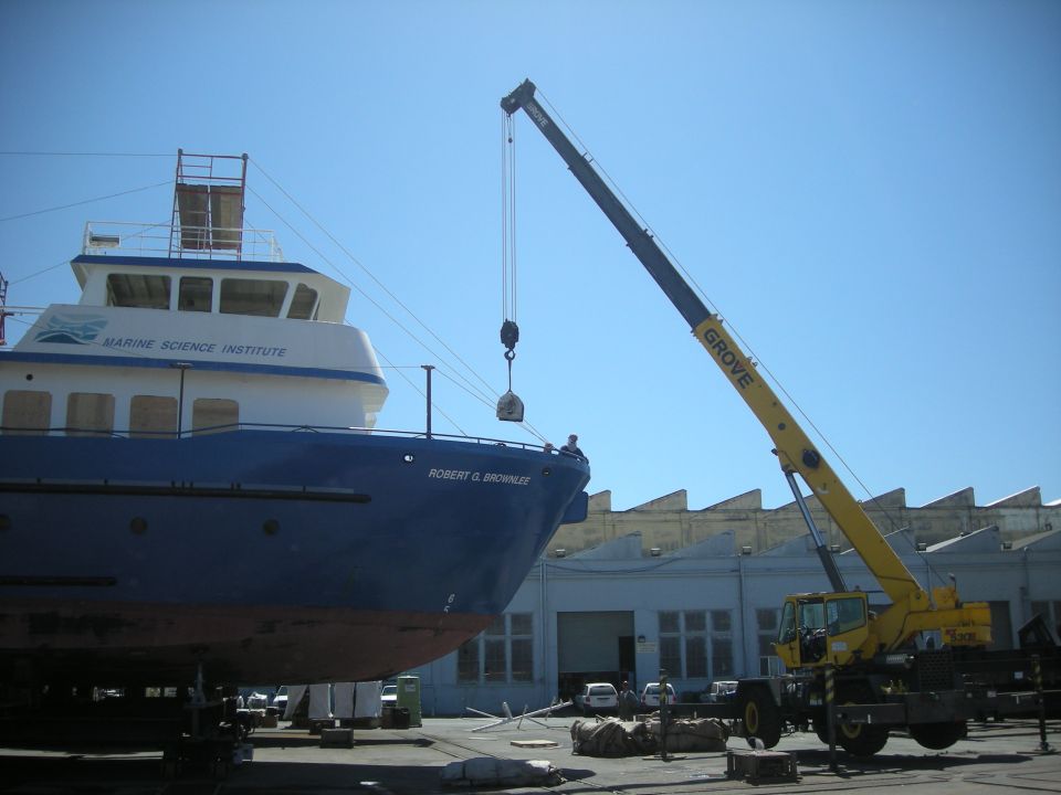 Blue and white boat named "ROBERT G. BROWNLIE" with "MARINE SCIENCE INSTITUTE" text, being lifted by a yellow crane