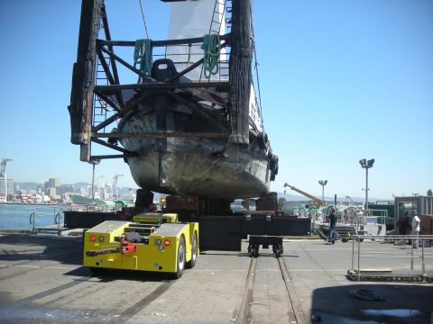 1991 Ford Bronco - A large boat is being transported on tracks by a yellow tugger vehicle in a sunny outdoor scene with a city skyline in the background.