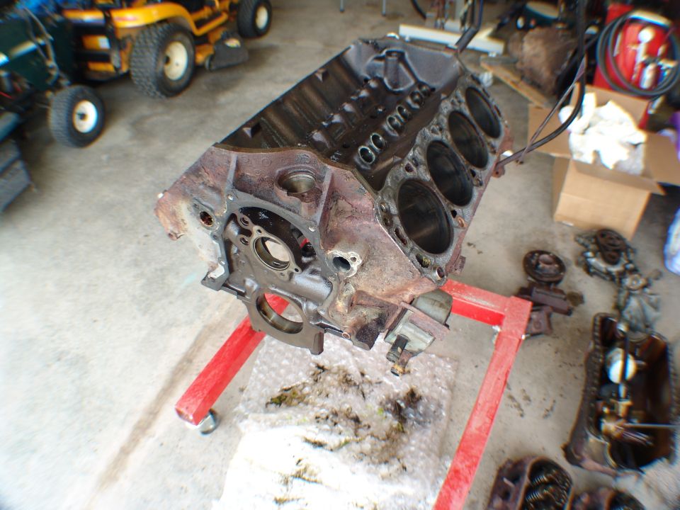 Stripped, rusty V8 engine block on a red engine stand in a garage with parts and tractors nearby