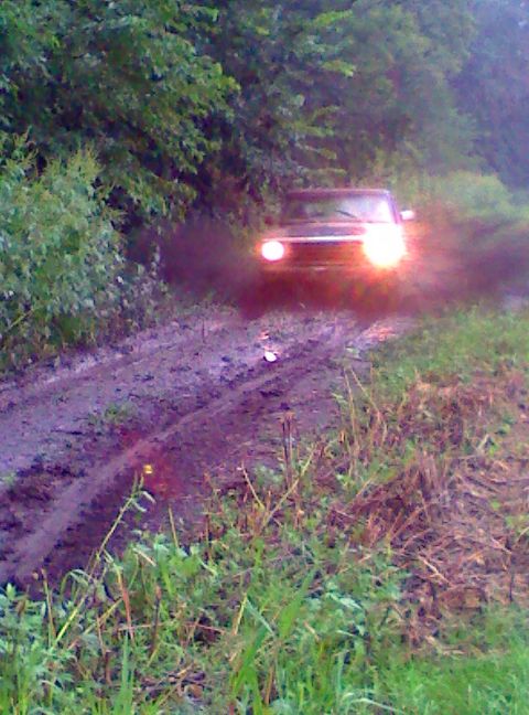 1979 Ford Bronco - A dark SUV navigates a muddy path, kicking up dirt as it drives with its headlights illuminated.