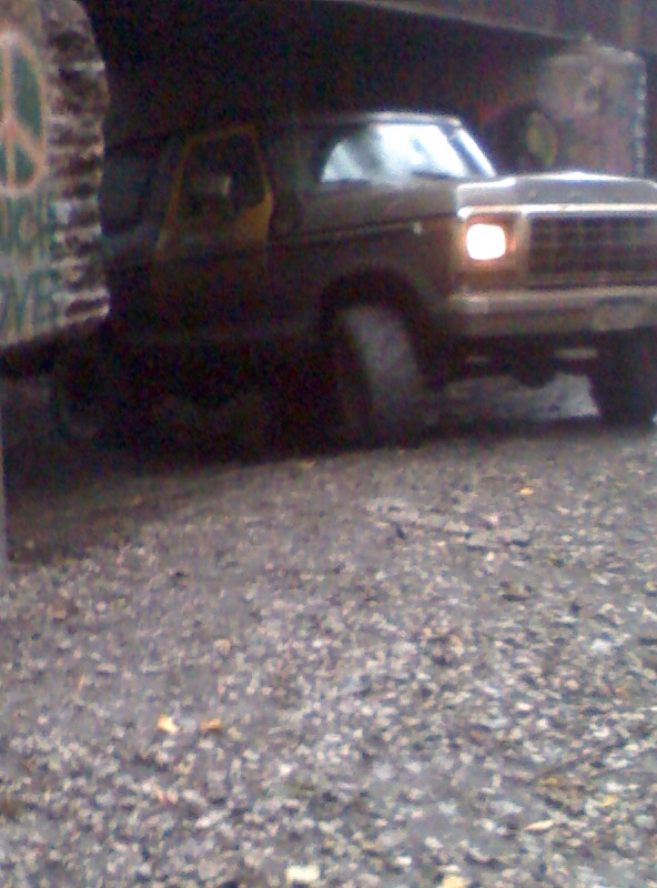 Dark brown 1978 Ford Bronco parked on gravel, front passenger side visible, headlight on